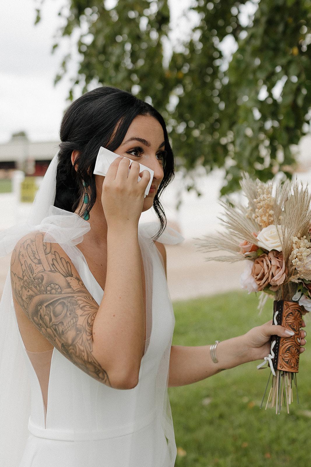 Bride dabbing tears with a tissue, tattoo sleeve and pampas bouquet on full display
