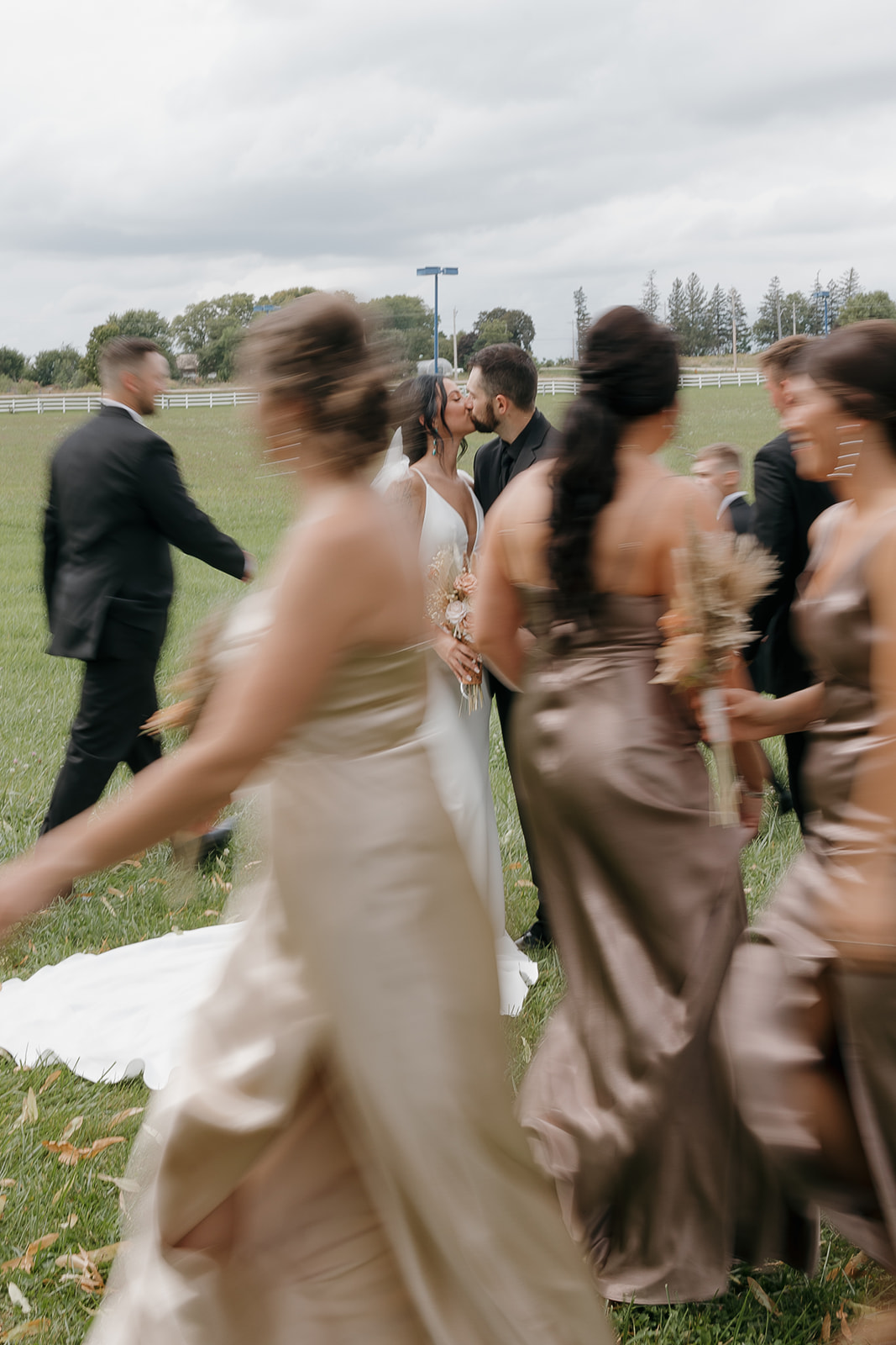 Motion blurred candid of bridesmaids walking past the bride and groom kissing in an open field