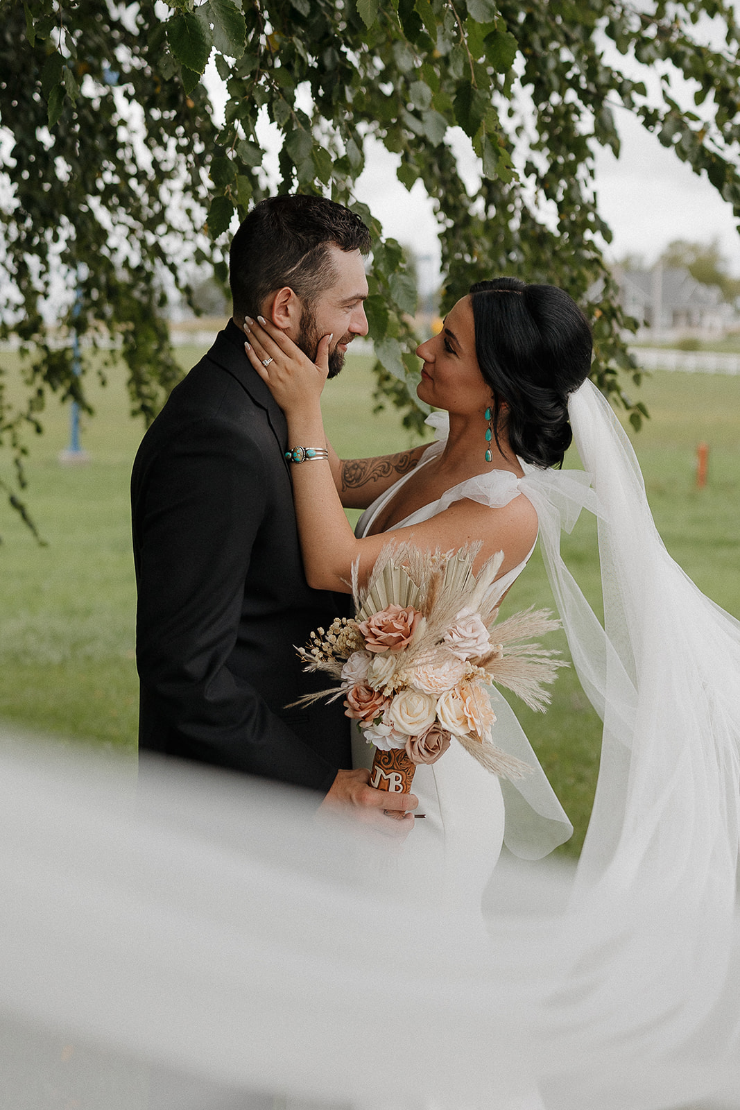 Bride and groom nose to nose under a tree, her hand on his cheek and veil flowing behind her