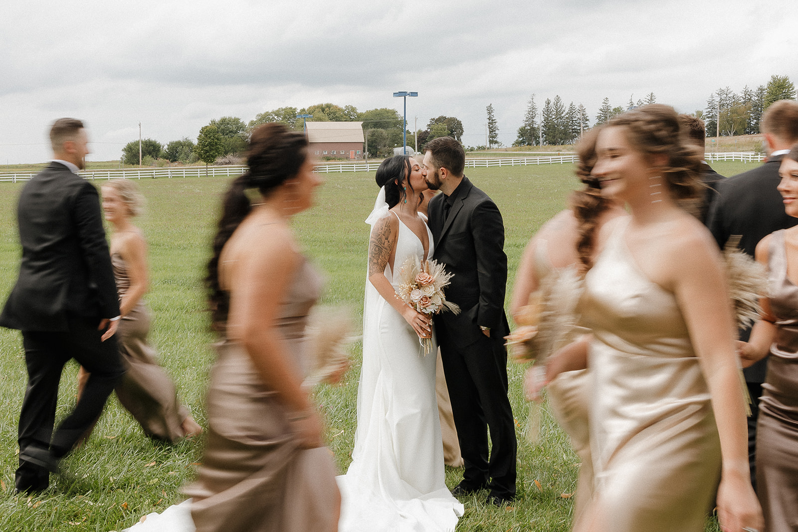 Candid motion shot of bride and groom kissing as bridal party walks past on a farm venue lawn
