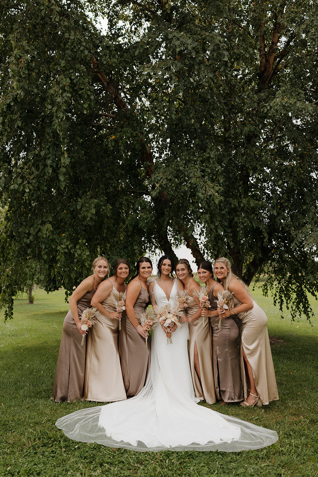 Bride and six bridesmaids in earth toned gowns posing together under a large tree
