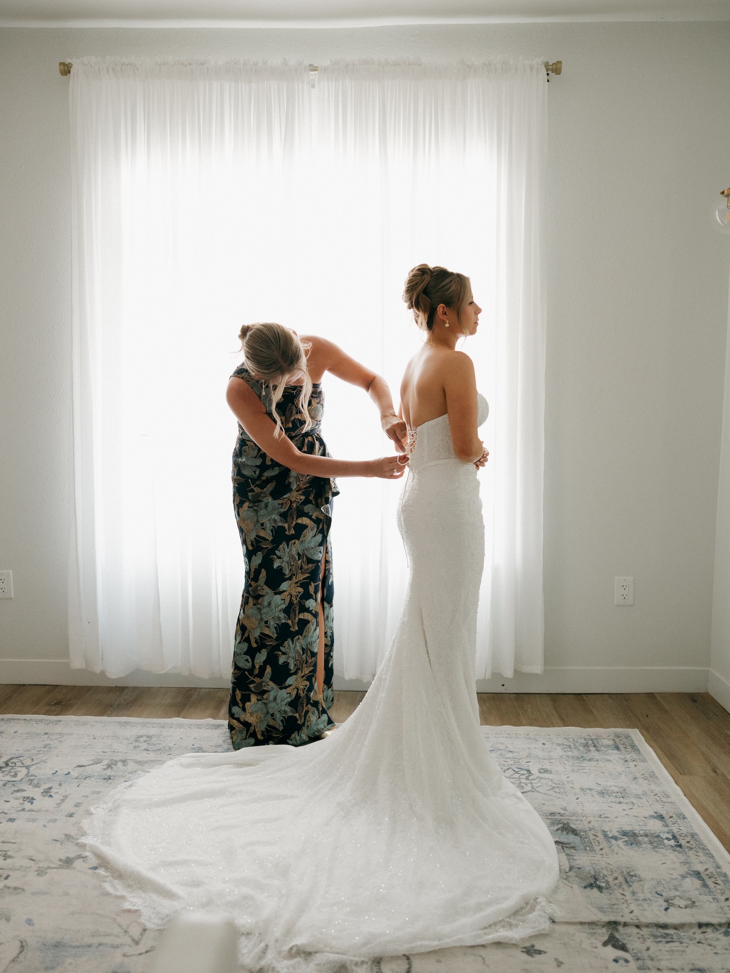 Bridesmaid lacing up the back of the bride's beaded wedding gown in a sunlit getting-ready room.