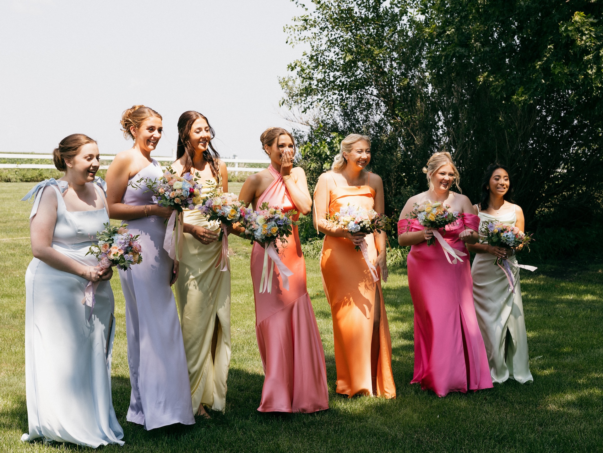 Bridesmaids in mismatched pastel gowns laughing together outdoors, each holding a wildflower bouquet.