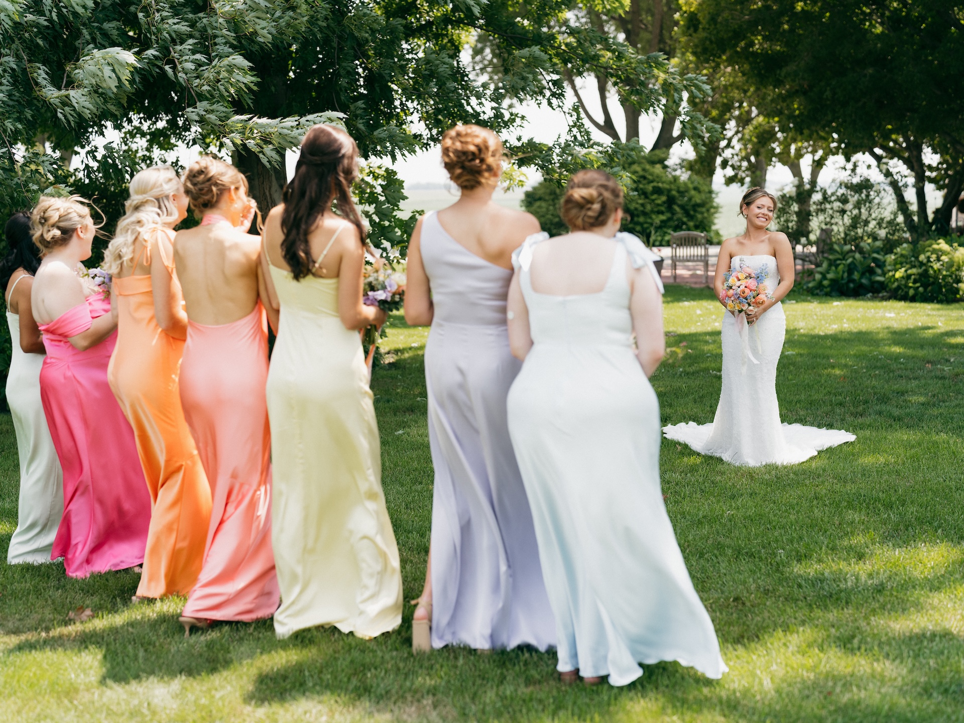 Bridesmaids in mismatched pastel gowns walking toward the bride waiting on the lawn before the ceremony.