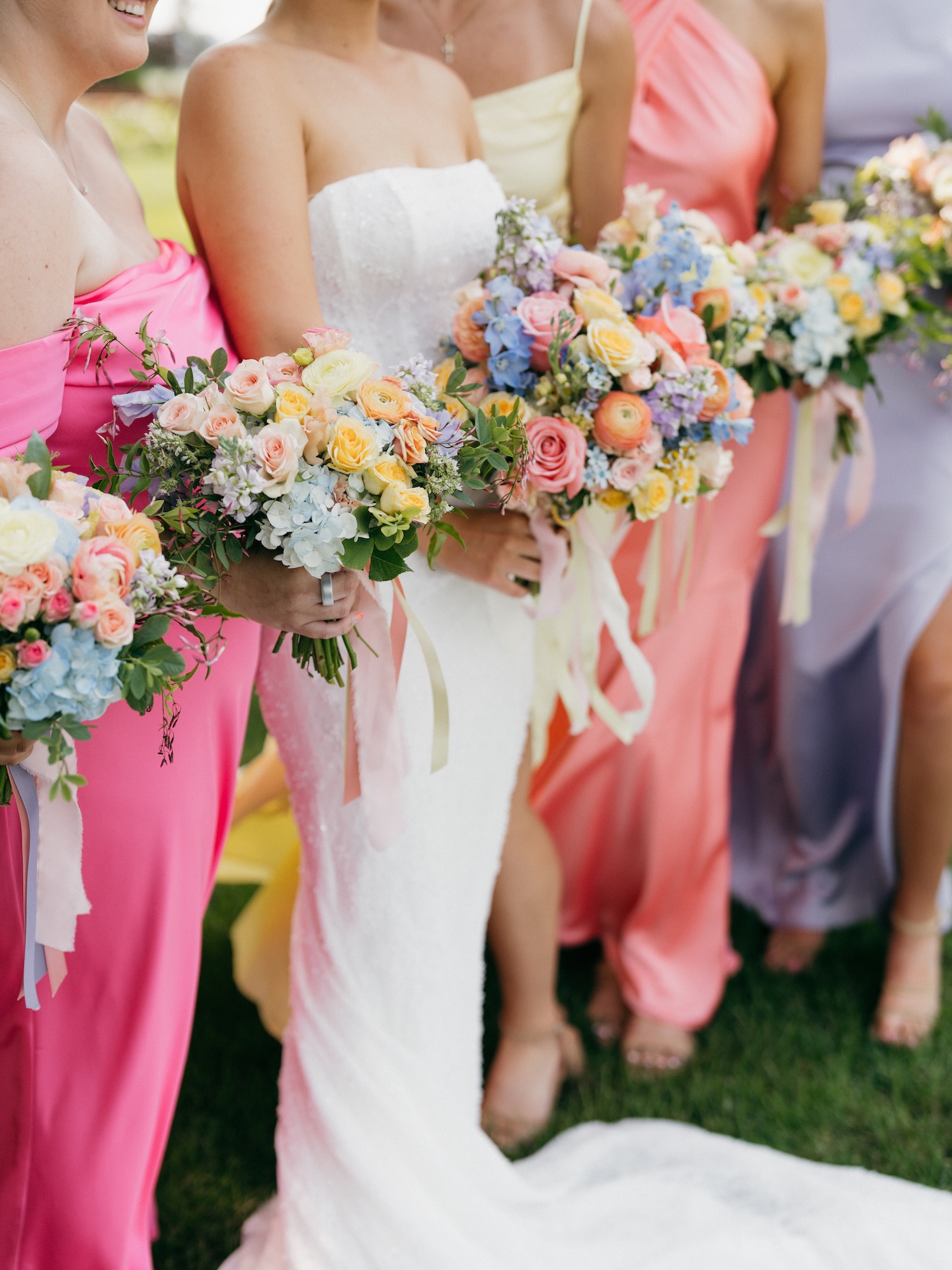 Close-up of the bride and bridesmaids' pastel bouquets of roses, delphinium, and ranunculus in a colorful wedding lineup.