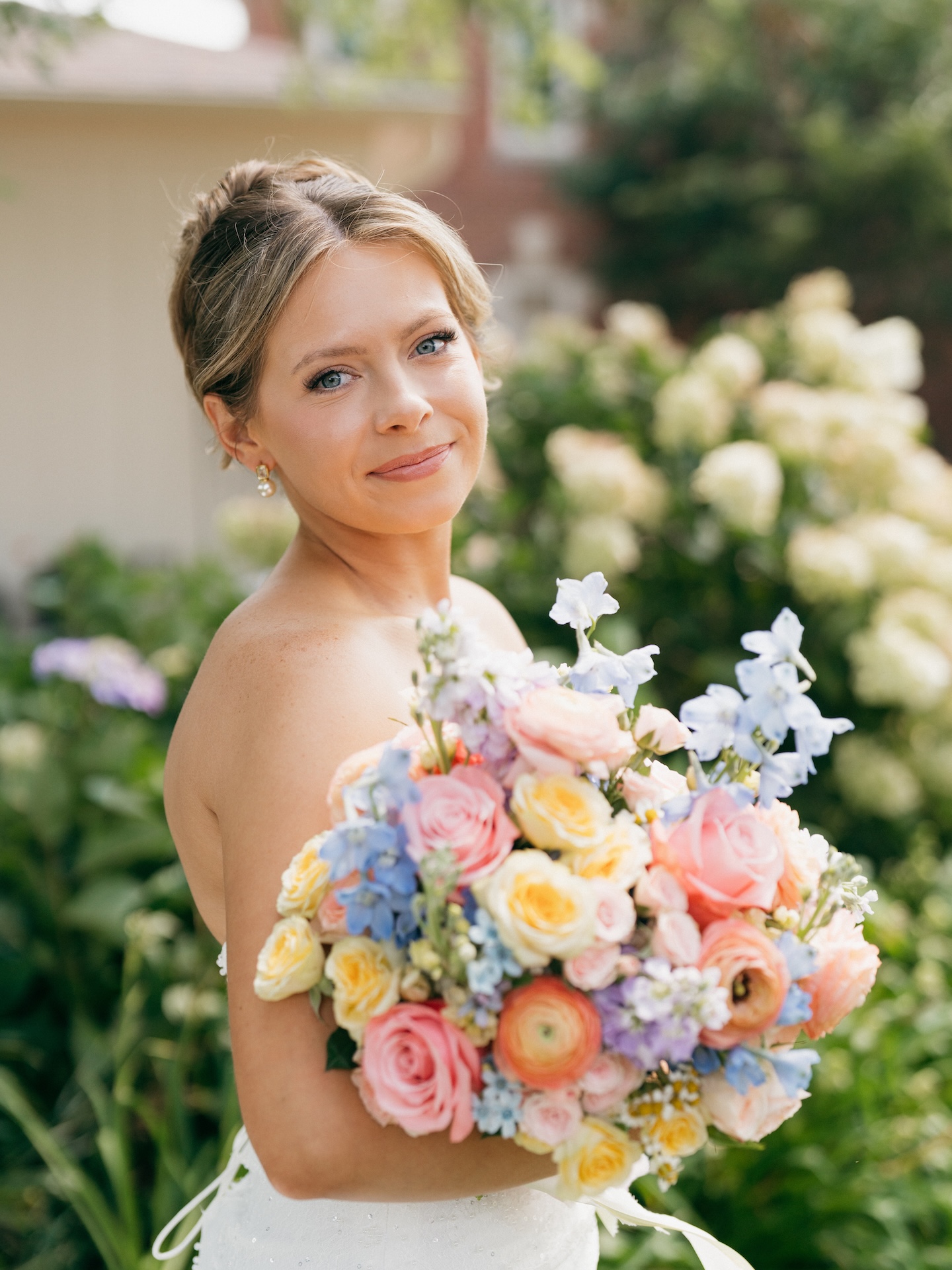 Bride in a strapless gown smiling over her shoulder while holding a colorful wedding bouquet of peach roses and blue delphinium.