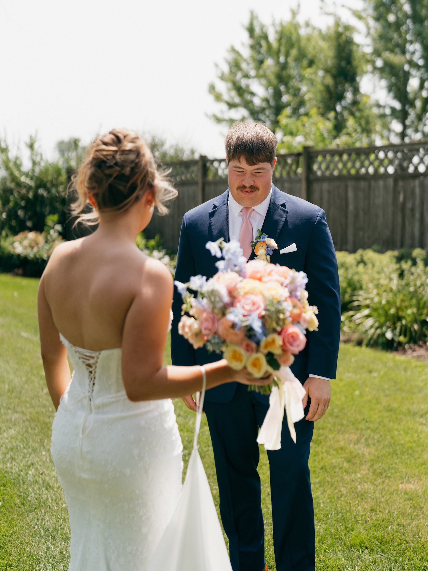 Bride showing her groom her bouquet of peach, blue, and yellow blooms during a backyard first look.