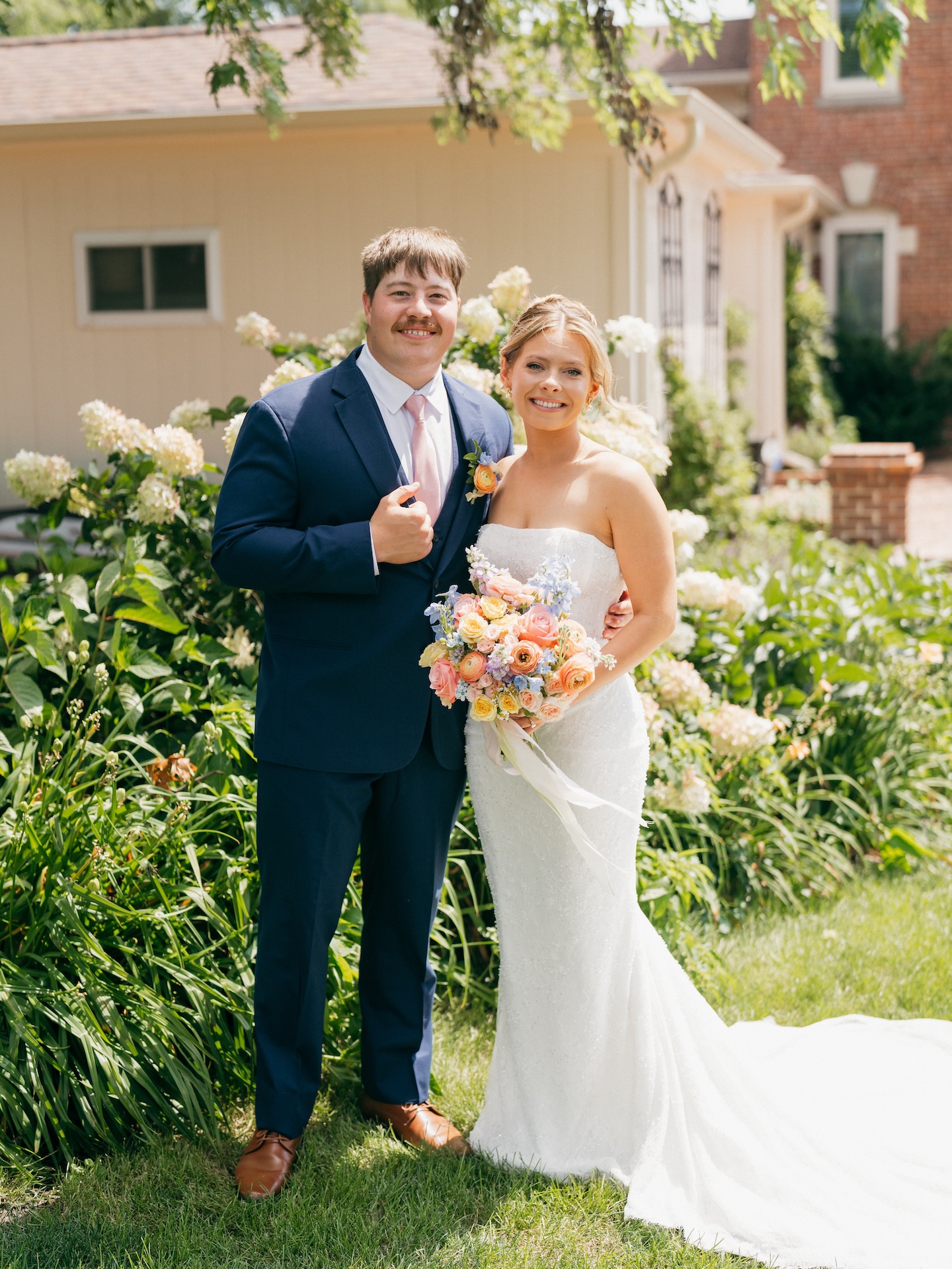Bride and groom smiling together in front of blooming white hydrangea bushes, colorful wedding bouquet in hand.