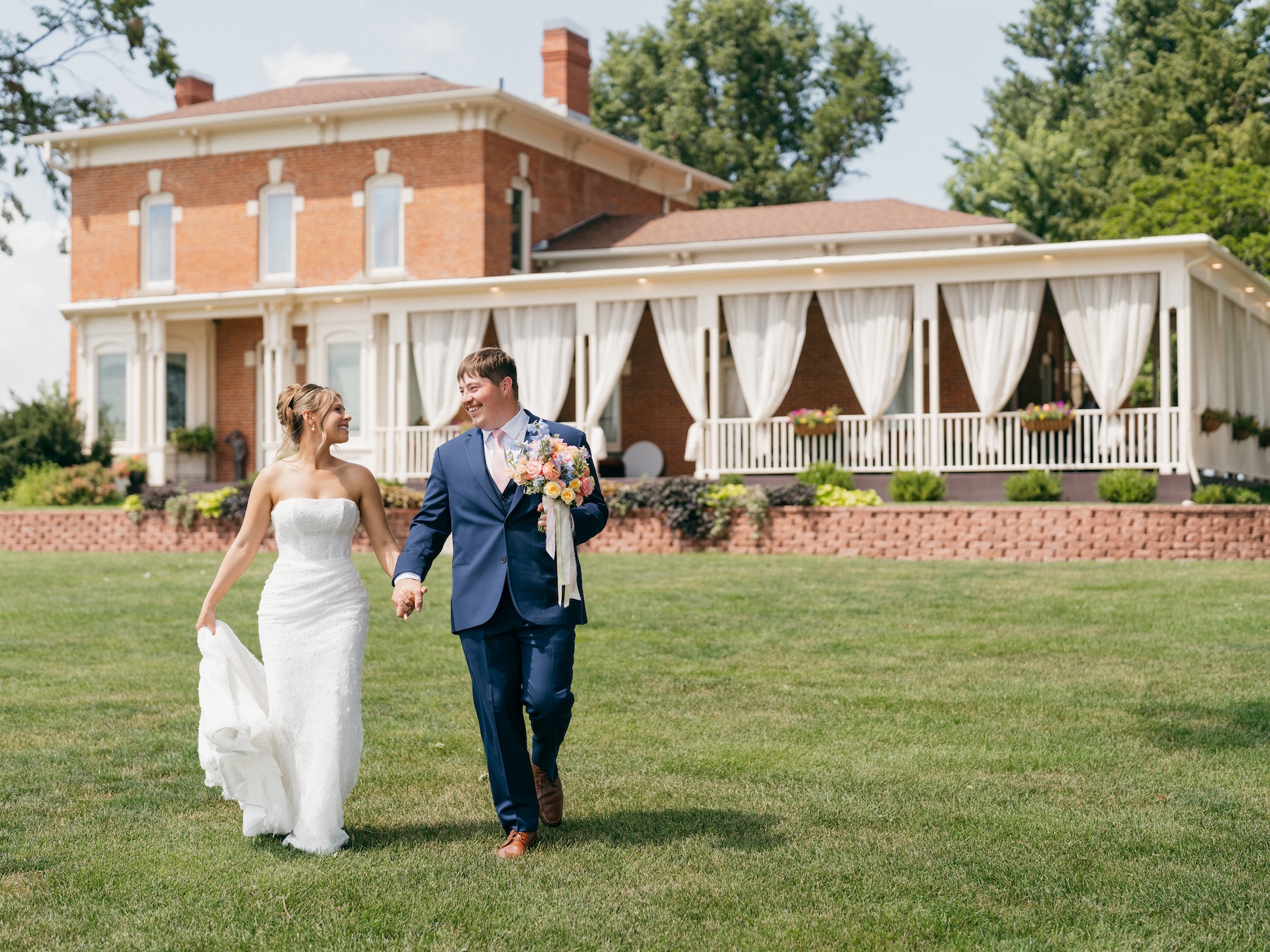 Bride and groom holding hands and laughing as they walk across the lawn in front of the brick farmhouse venue.