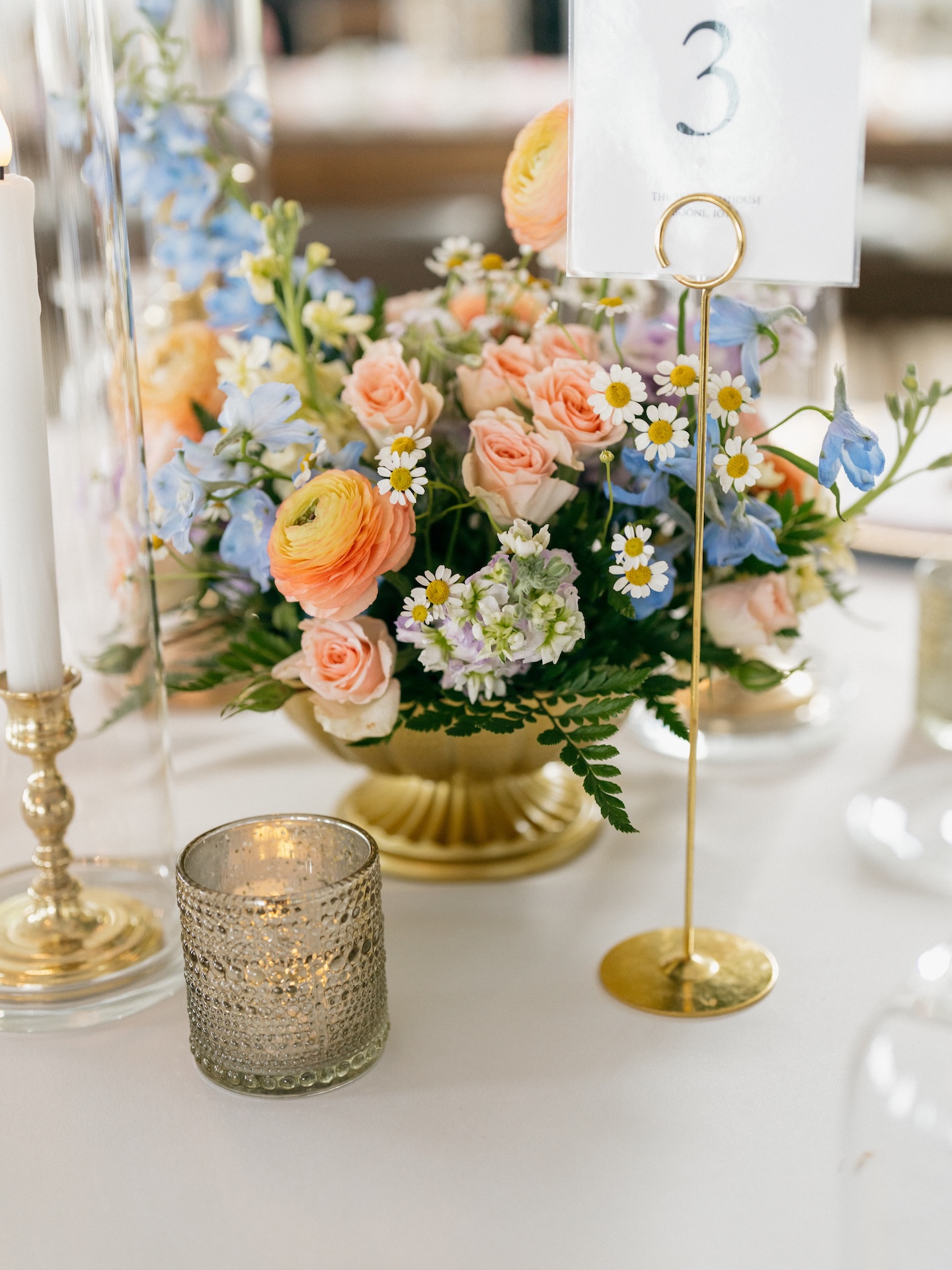 Gold compote centerpiece with peach roses, blue delphinium, and daisies beside a gold table number stand, colorful wedding reception decor.