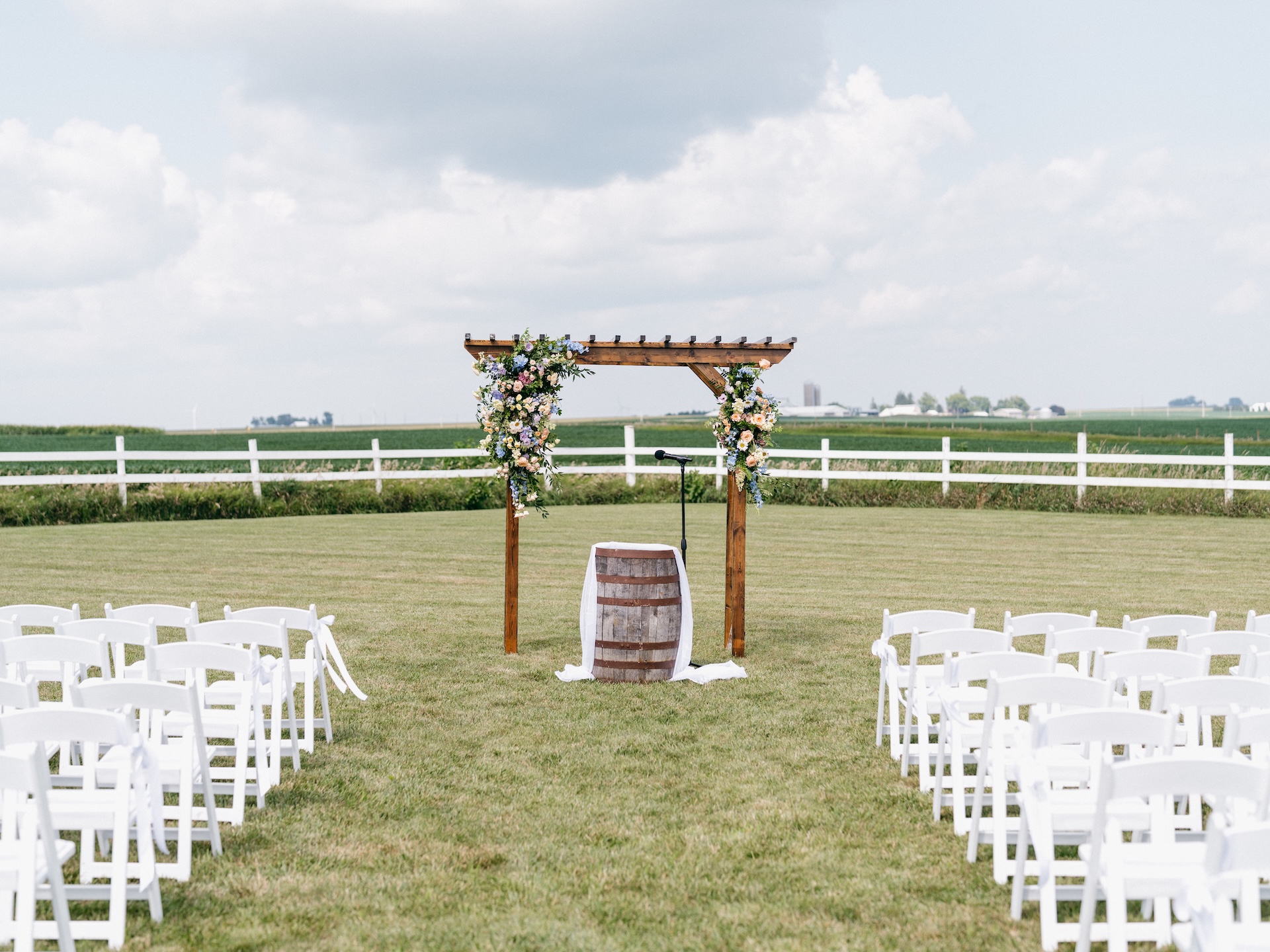Floral wedding arbor and empty white chairs set up in a farm field before the ceremony begins.
