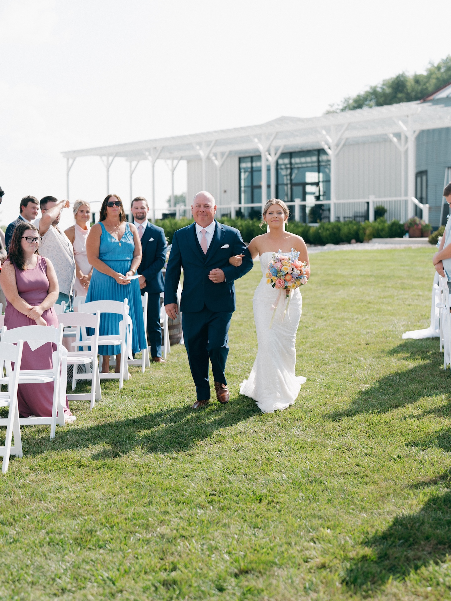 Bride walking down the aisle arm in arm with her father past guests seated in white folding chairs.