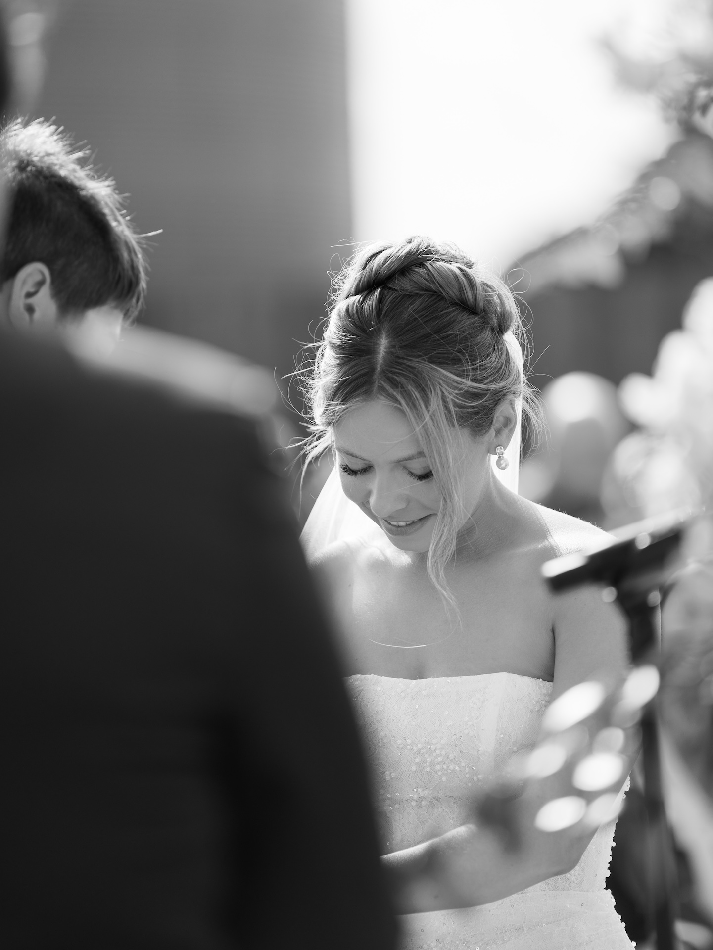 Black and white candid of the bride smiling during her wedding ceremony as the groom looks on.
