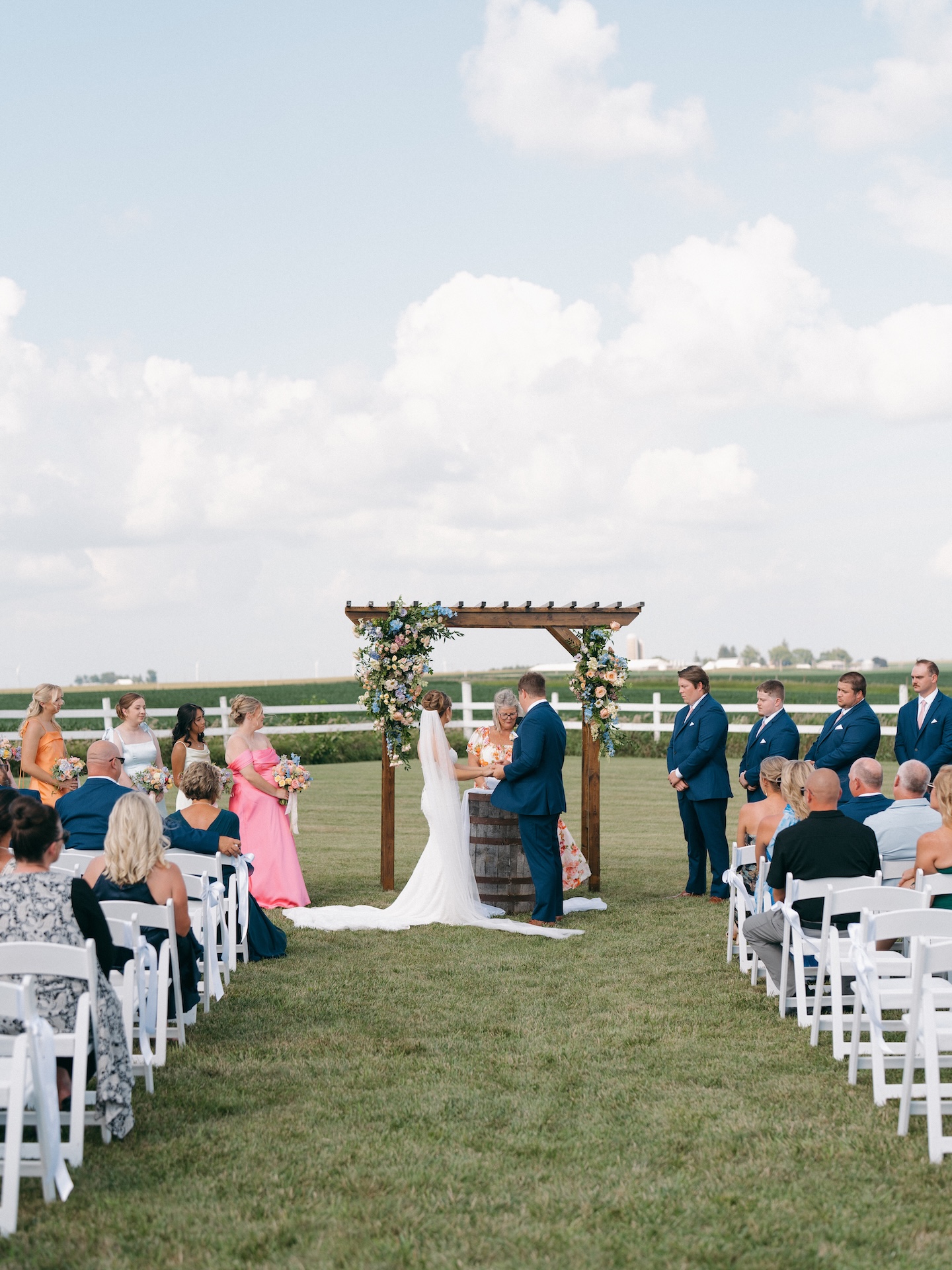 Wide shot of the outdoor farm ceremony with guests seated in white chairs facing the flower-covered arbor, colorful wedding florals in bloom.