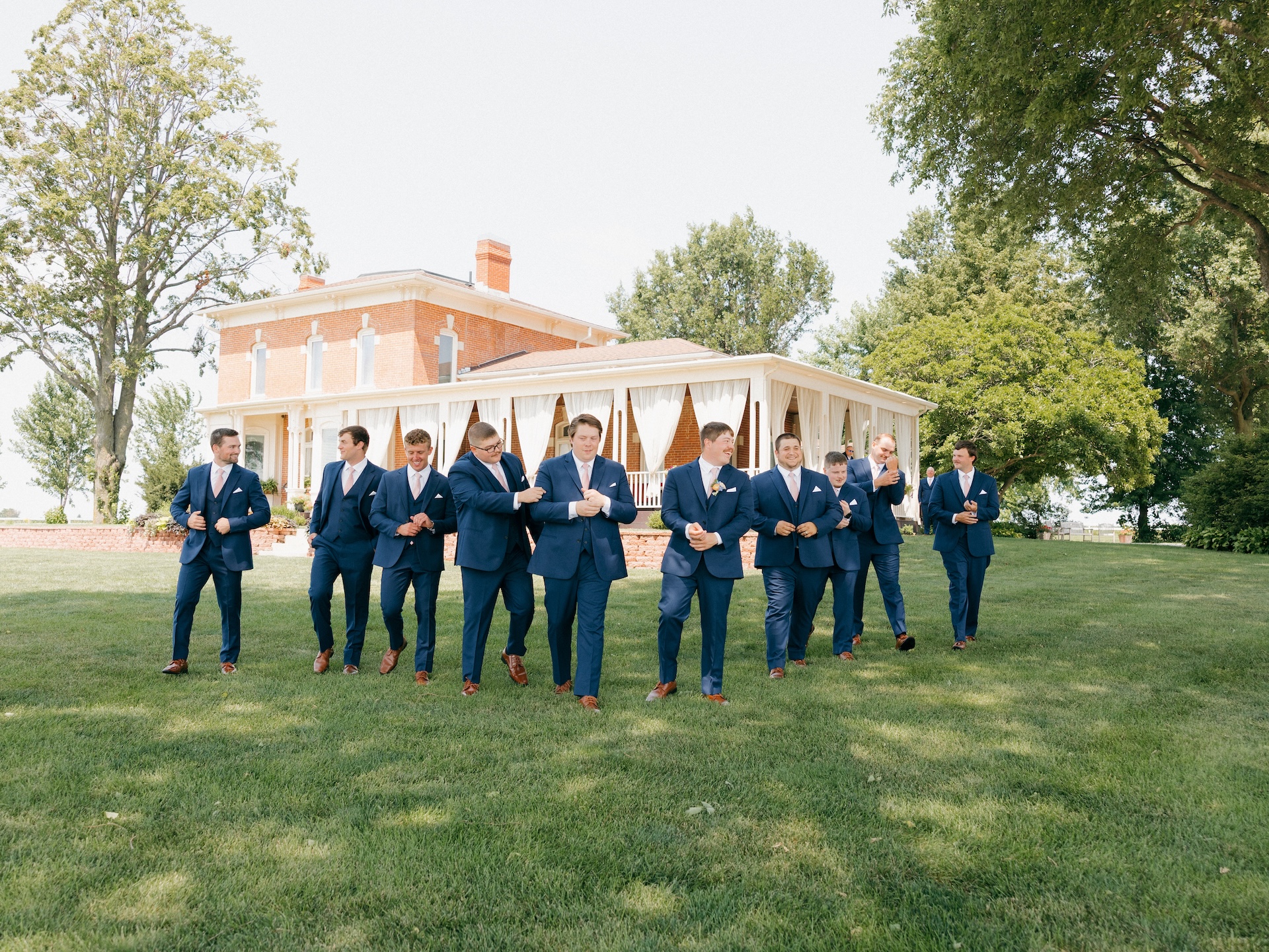 Groomsmen in matching navy suits and pink ties walking across the lawn in front of the brick farmhouse venue, colorful wedding party portraits.