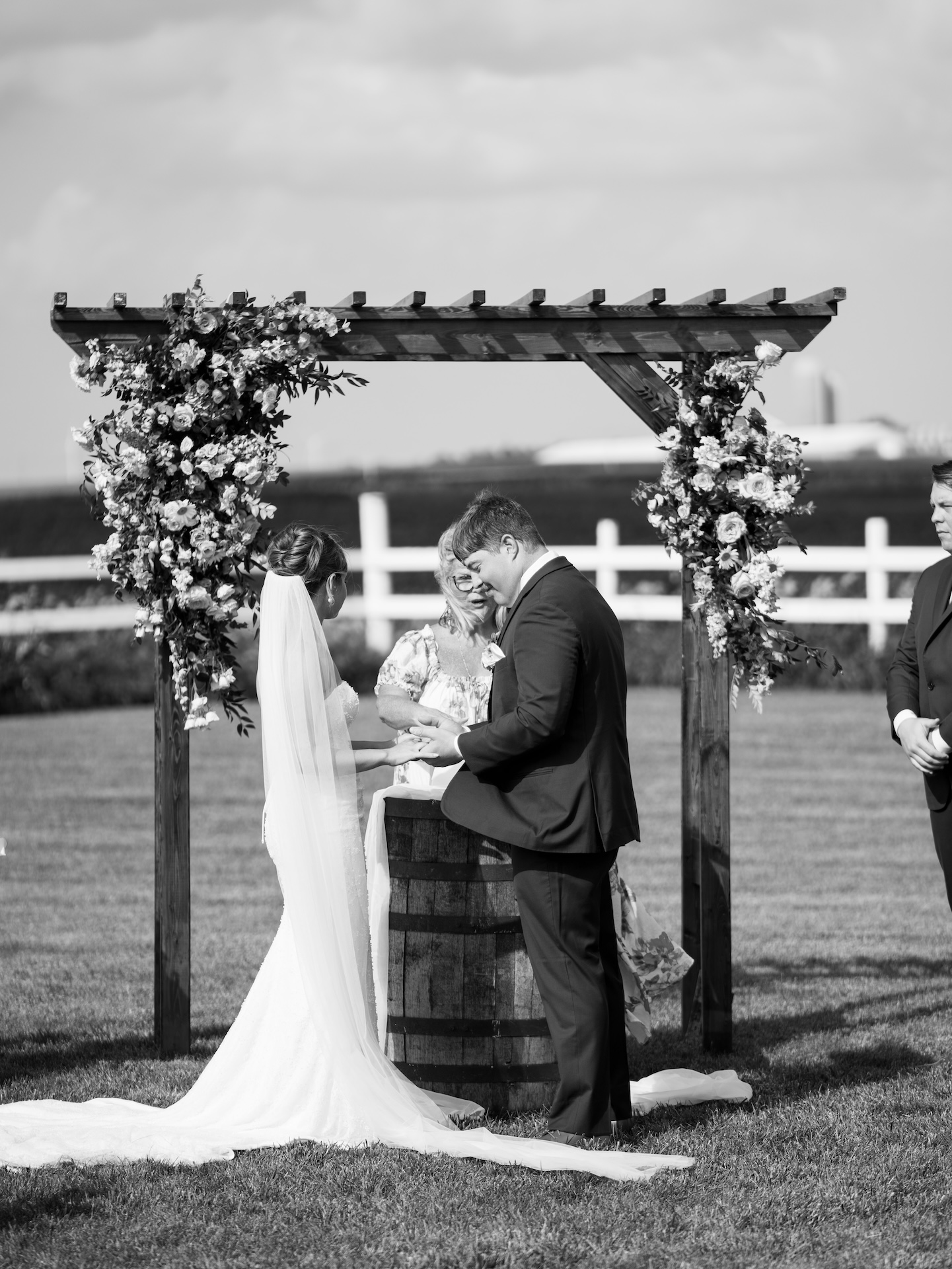 Black and white photo of the bride and groom holding hands during their vows beneath a flower-covered arbor.