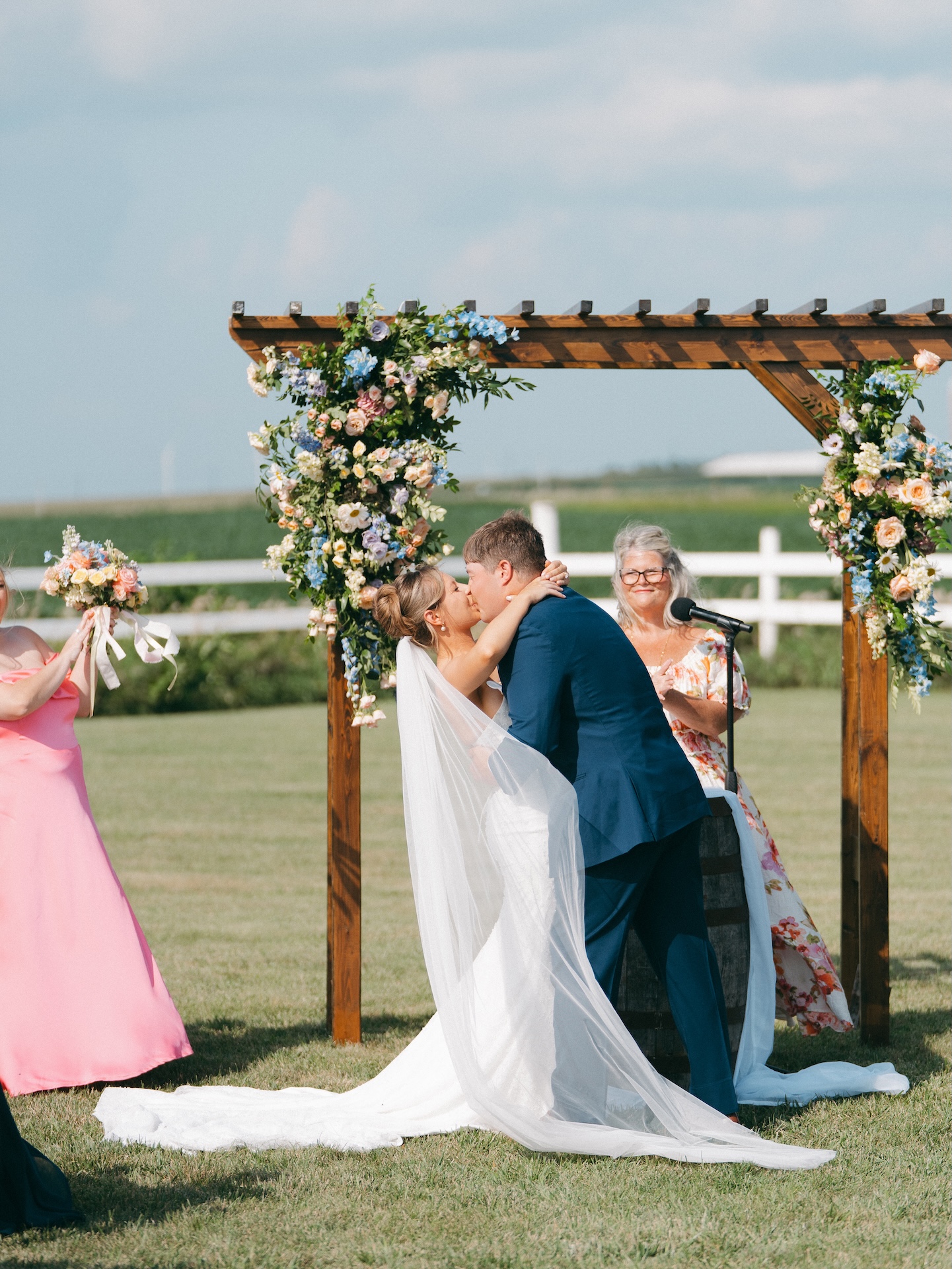 Bride and groom sharing their first kiss as a married couple beneath the flower-covered arbor, colorful wedding guests looking on.