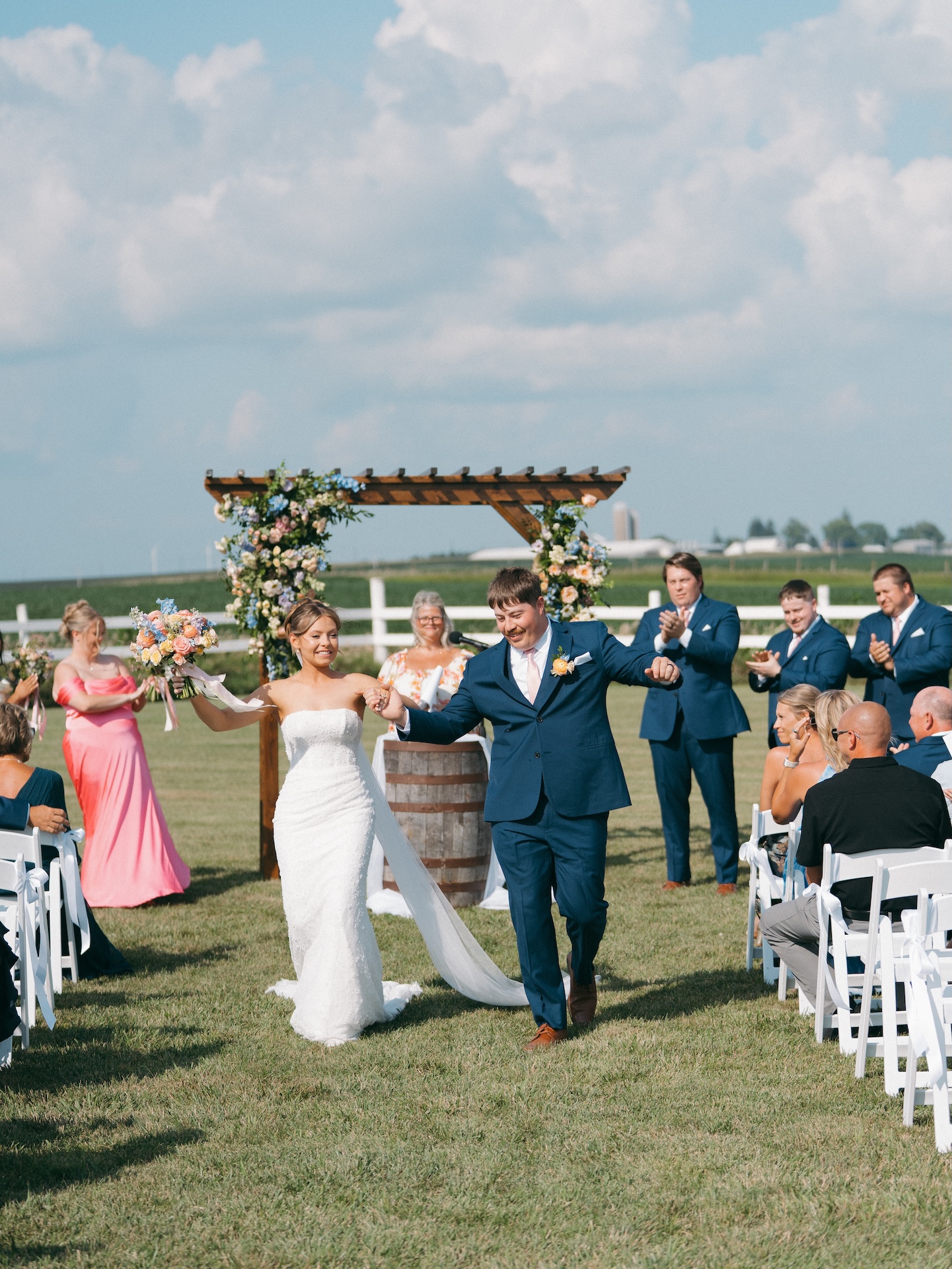 Newlyweds walking back up the aisle hand in hand as guests applaud beneath the floral ceremony arbor.