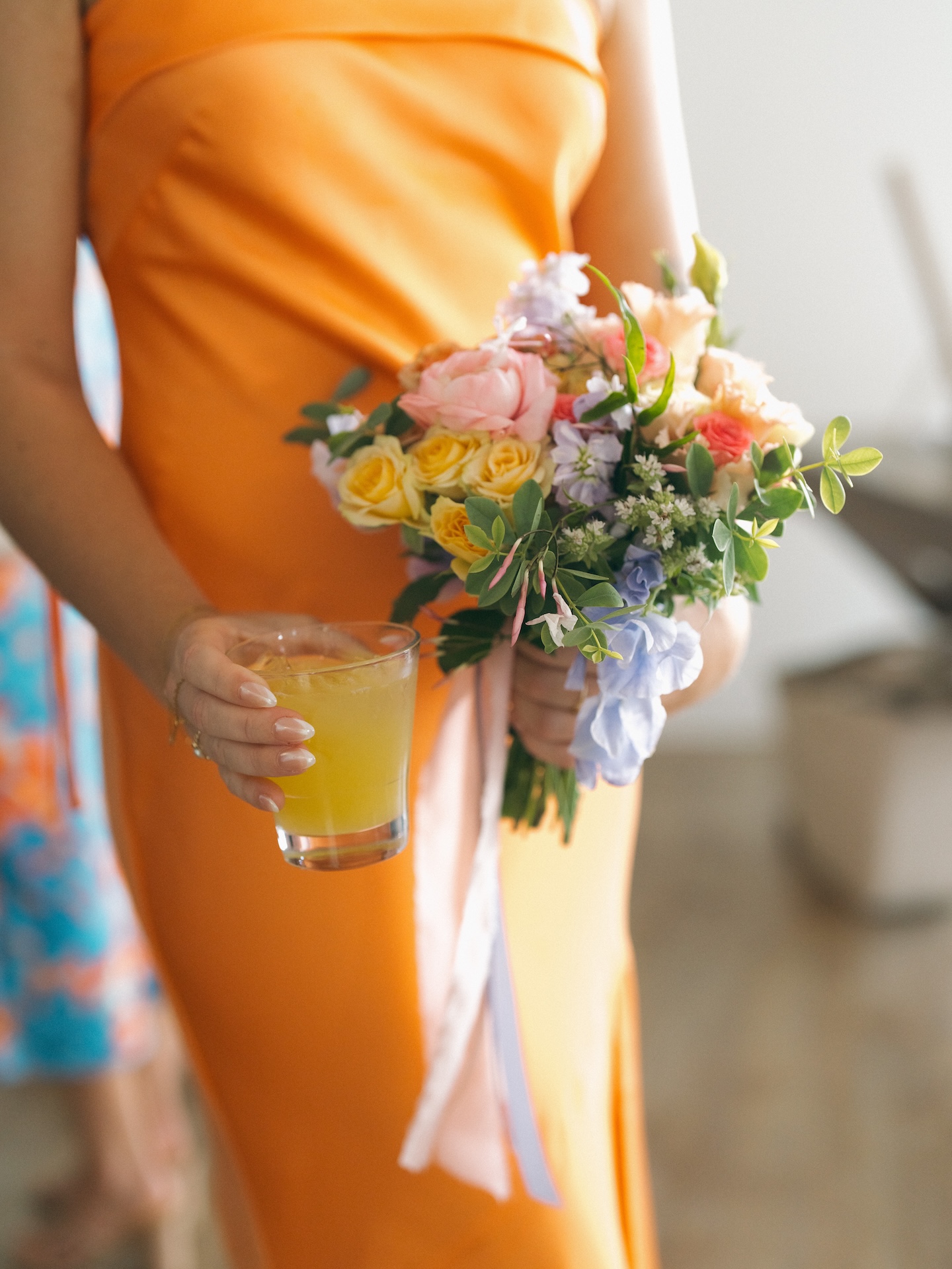 A bridesmaids holding a cocktail and her bouquet in front of her bright orange bridesmaid dress.
