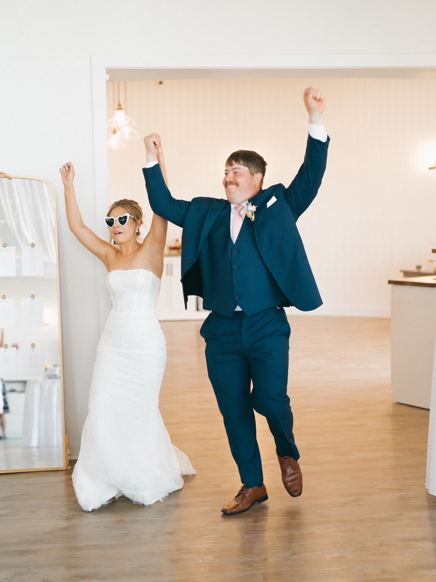Bride in heart-shaped sunglasses and groom throwing their arms up in celebration at the reception entrance.