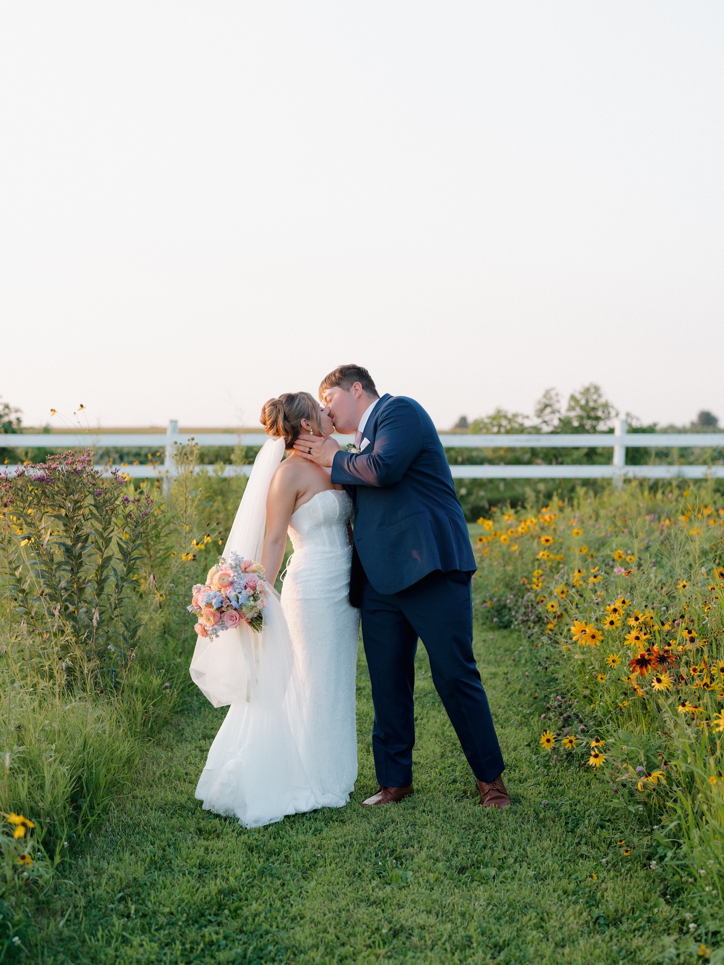 Groom holding his bride close as they laugh together in front of the brick farmhouse at sunset.