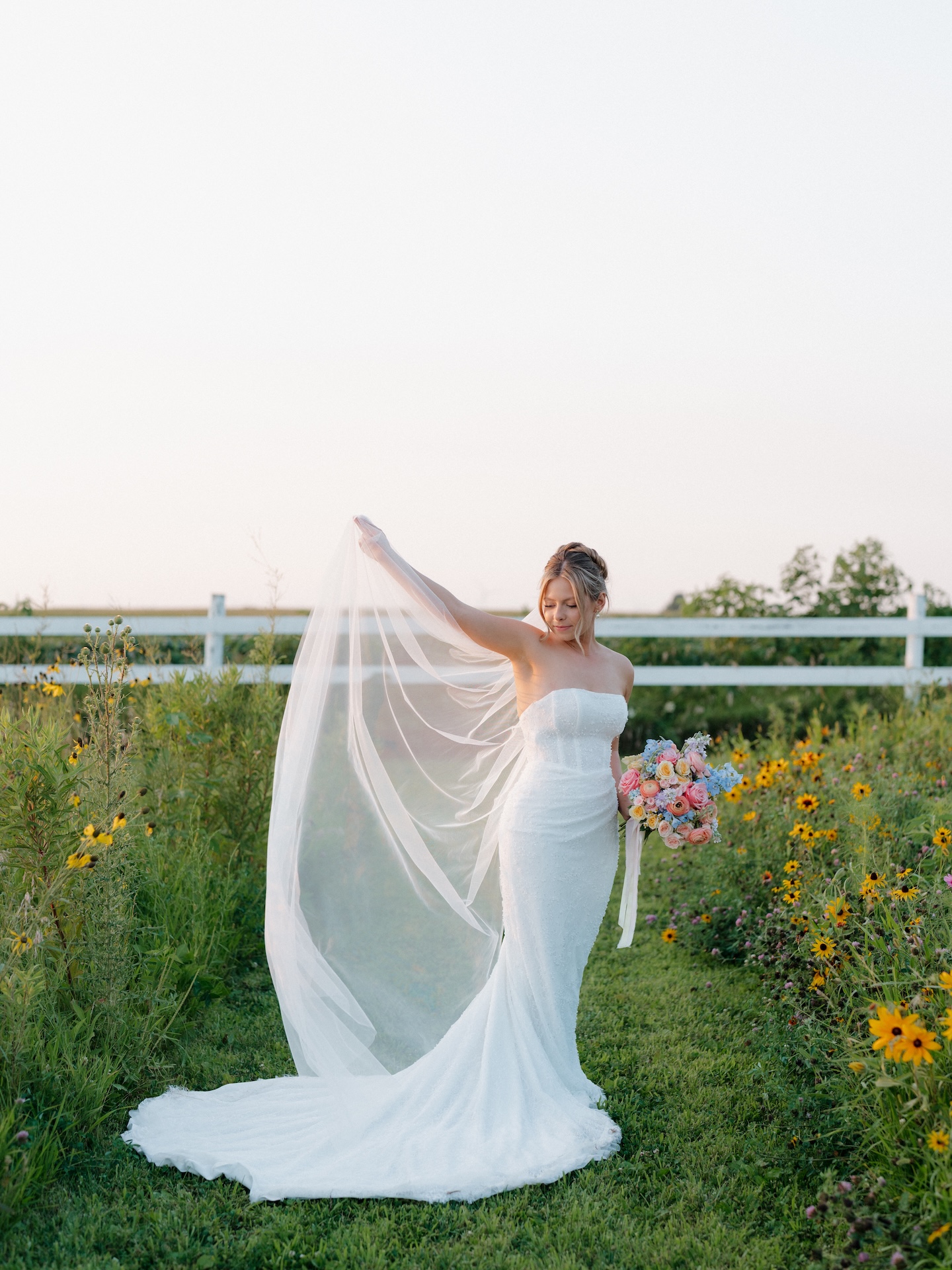 Bride holding her veil aloft in a wildflower field, colorful wedding bouquet of roses and delphinium in hand.