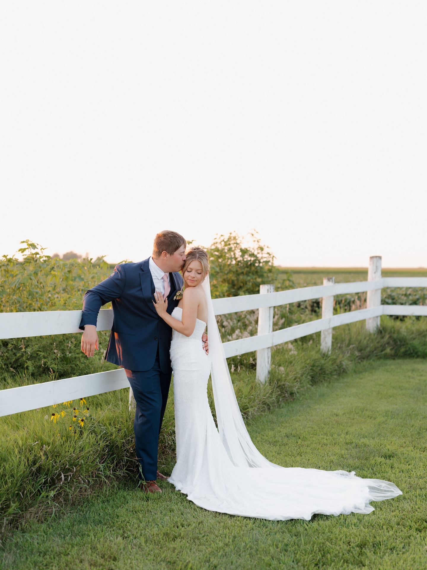 Groom kissing his bride's forehead beside a white farm fence at golden hour after their colorful wedding.