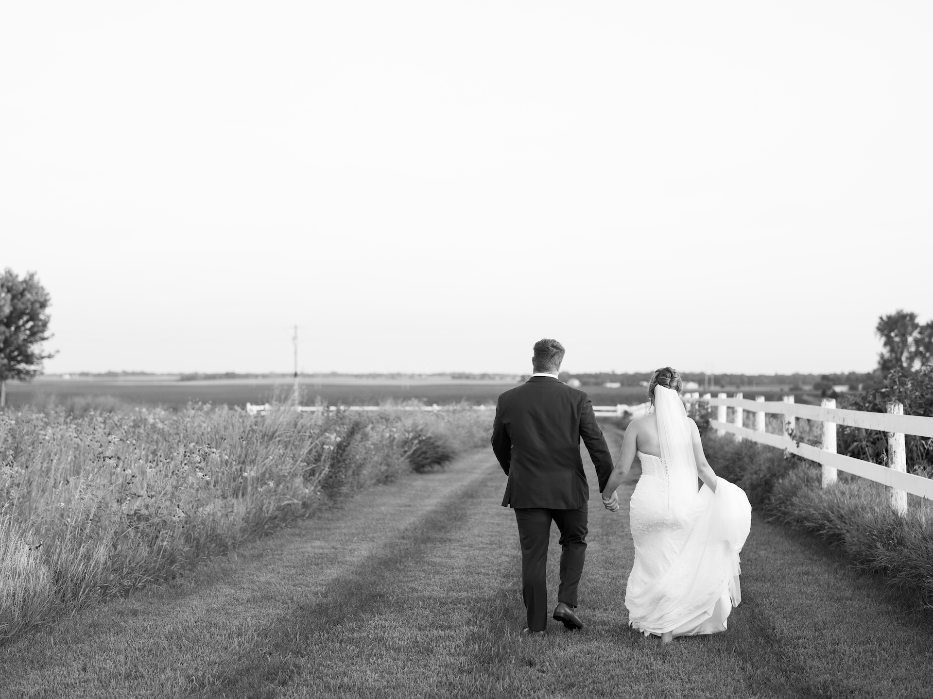 Black and white photo of the bride and groom walking hand in hand down a farm lane after their wedding.
