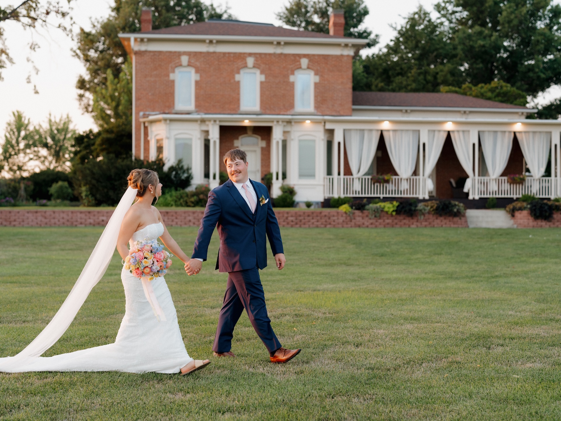 Bride and groom walking hand in hand across the lawn in front of the brick 1868 Farmhouse venue at golden hour.