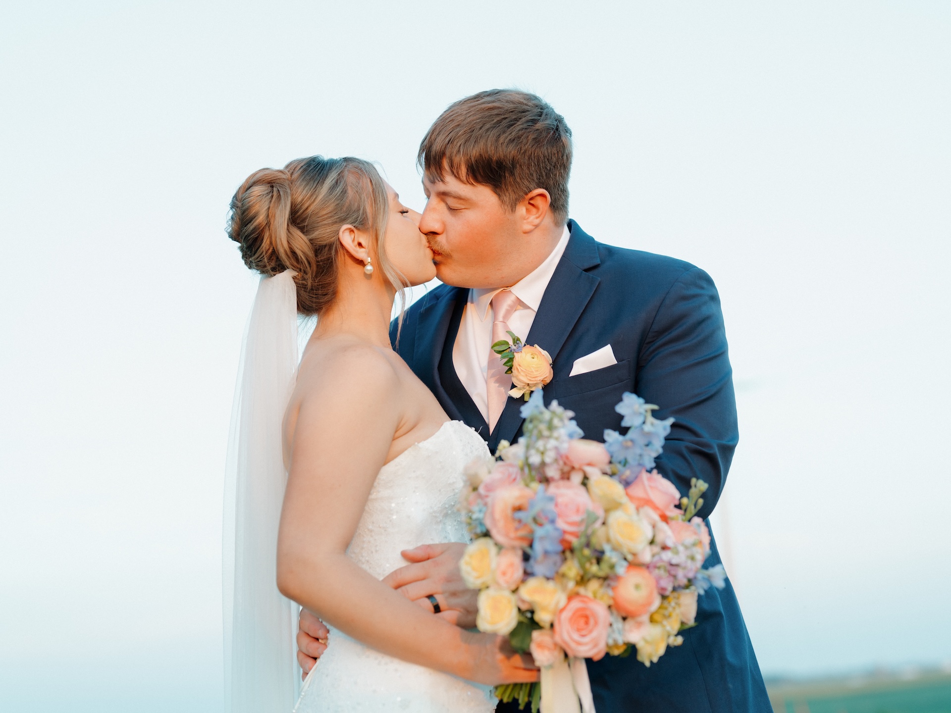Bride and groom sharing a kiss against an open sky, colorful wedding bouquet of peach roses and blue delphinium in view.