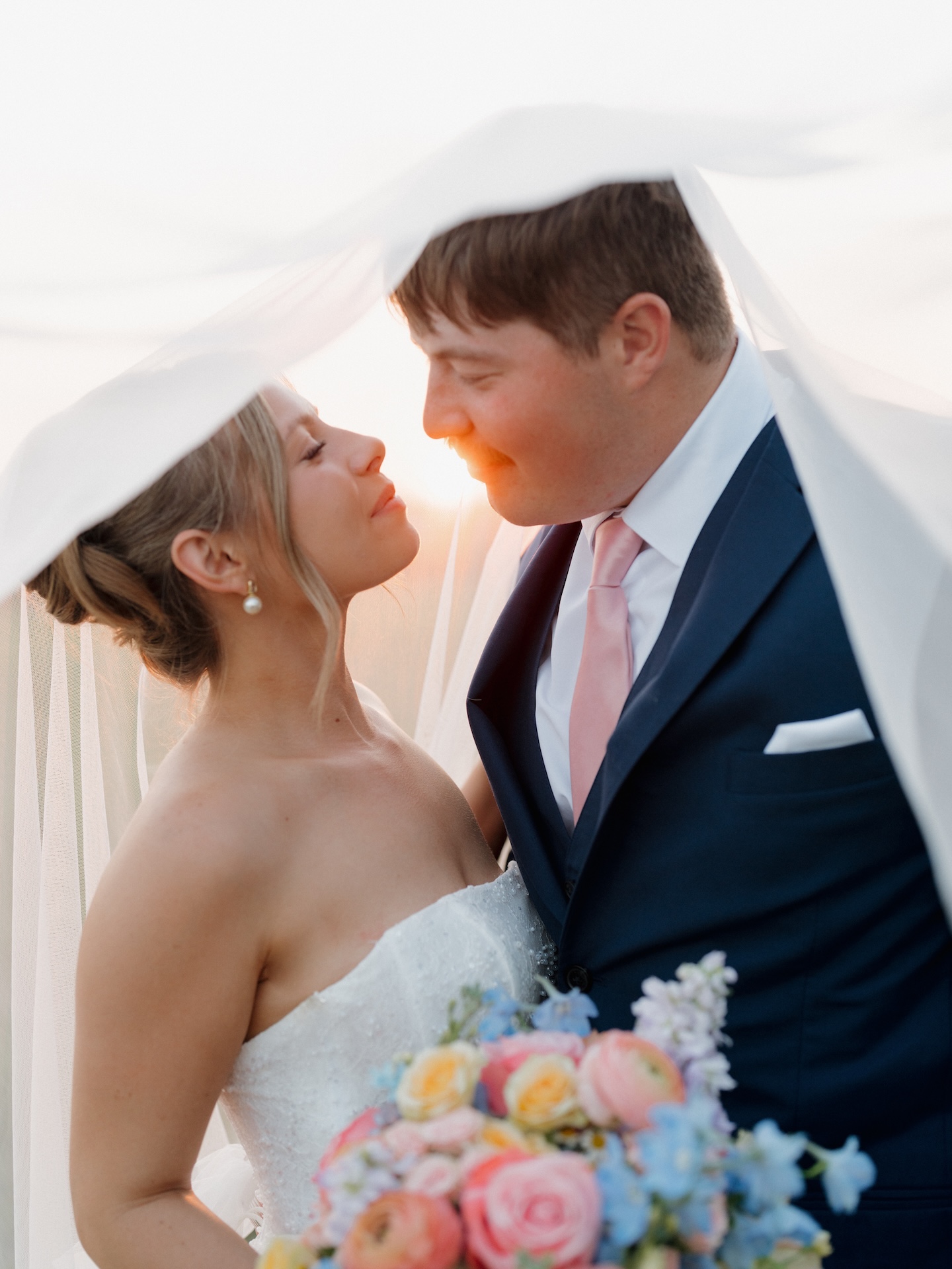 Bride and groom kissing beneath a flowing veil backlit by the setting sun, colorful wedding bouquet in view.
