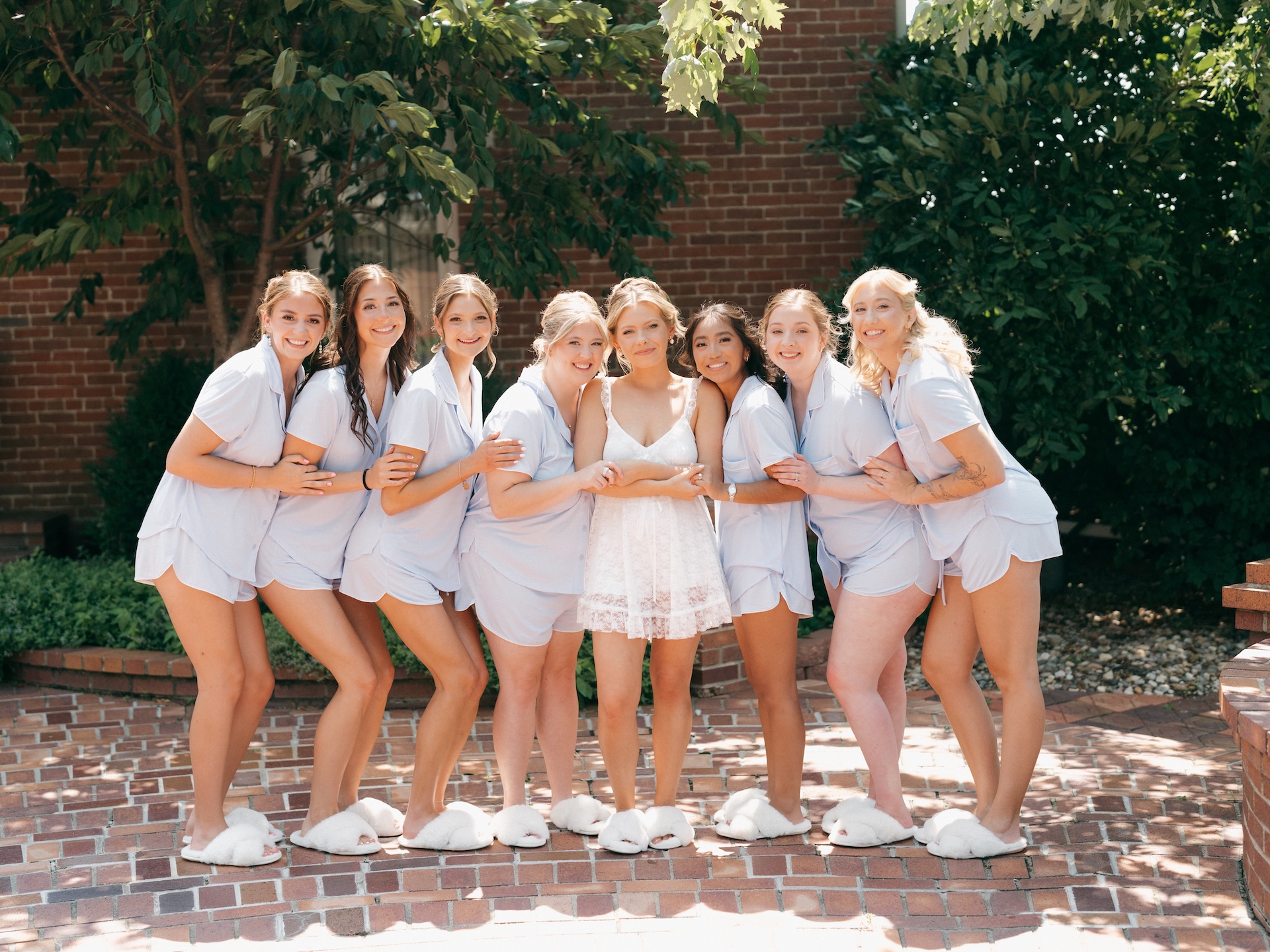 Bridesmaids in mismatched pastel jumpsuits and gowns smiling together on the lawn, bouquets in hand.