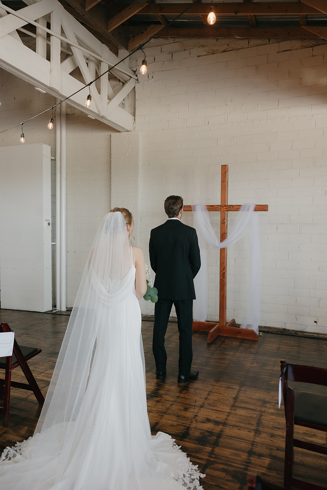 Bride and groom getting ready for the first look facing a wooden cross draped in tulle, a quiet moment before their ceremony at this modern wedding venue (Highpoint Event Center).