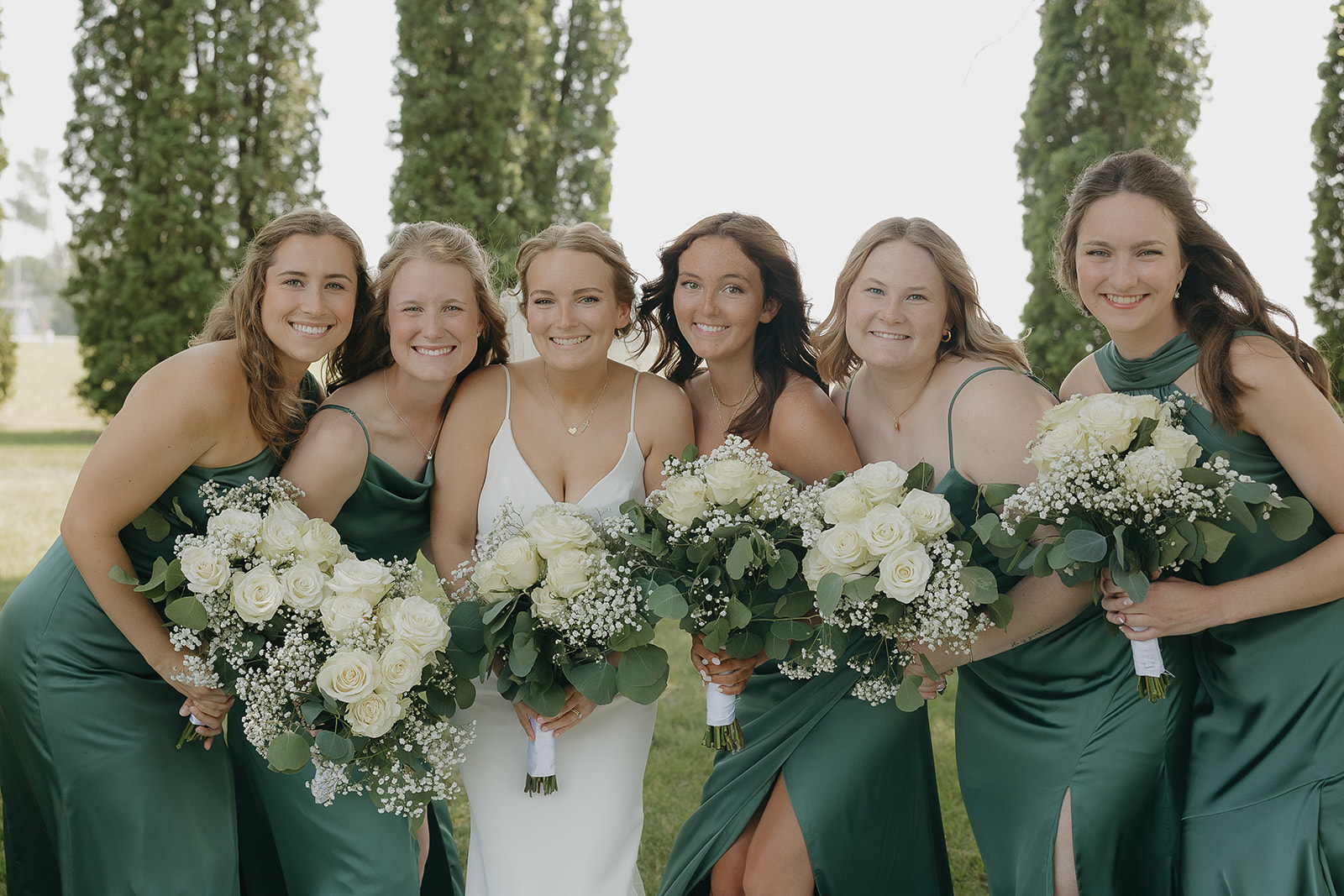 Bride surrounded by her bridesmaids in emerald green dresses, all holding white rose bouquets.