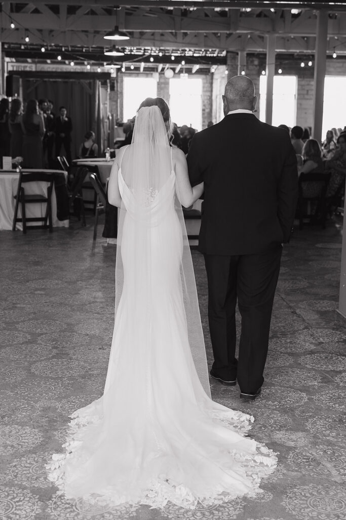 Black and white photo of the bride and her father walking arm in arm through the reception space at this modern wedding venue (Highpoint Event Center).