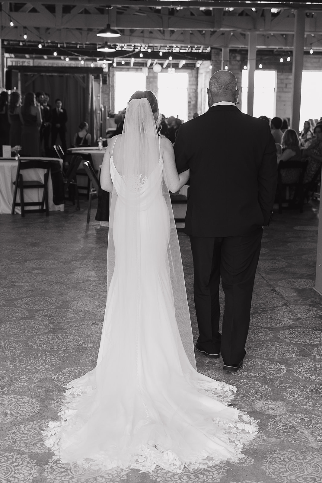 Black and white photo of the bride and her father walking arm in arm through the reception space at this modern wedding venue (Highpoint Event Center).
