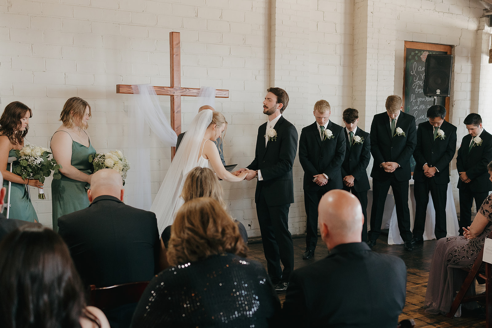 Bride and groom holding hands during their vows with the wedding party standing behind them.