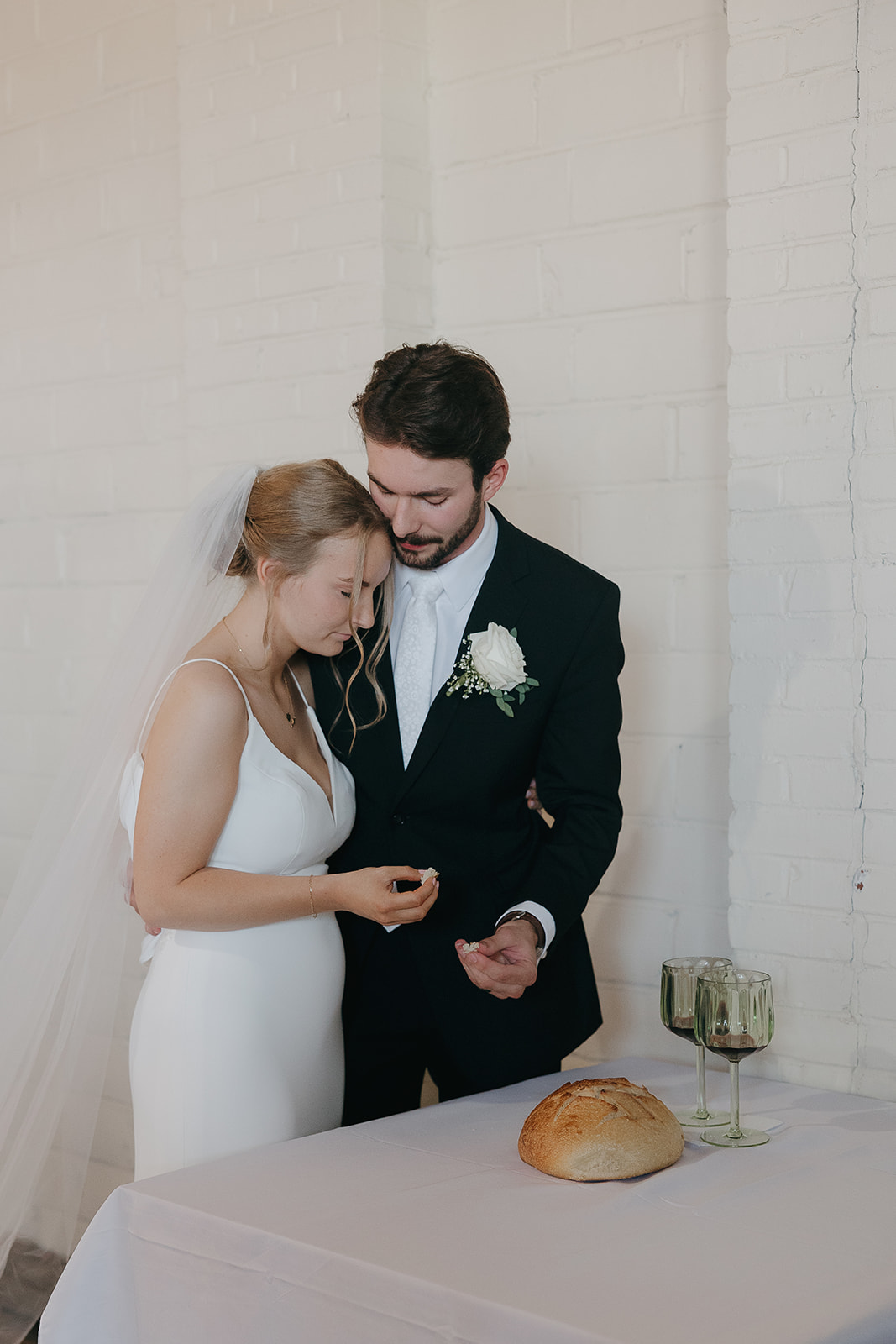 Bride and groom sharing a quiet communion moment with bread and wine set on a table at this modern wedding venue (Highpoint Event Center).