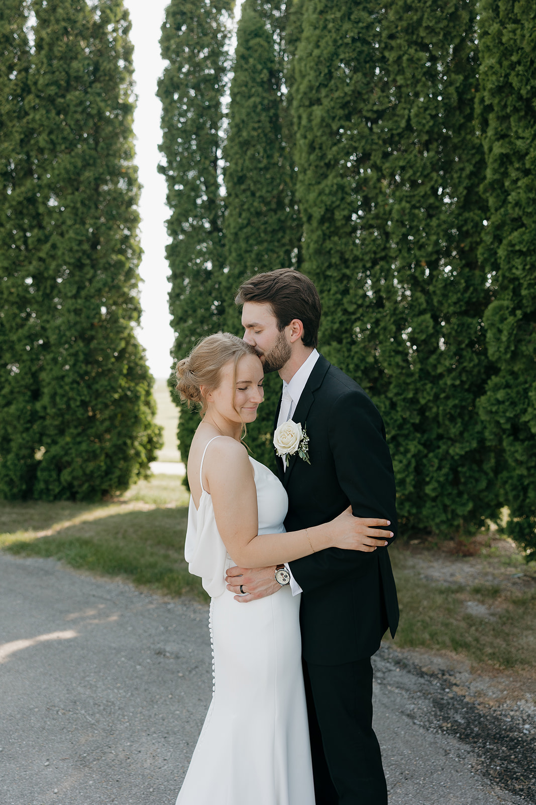 Groom kissing his bride's forehead along a tree lined pathway, a romantic portrait moment at this modern wedding venue (Highpoint Event Center).