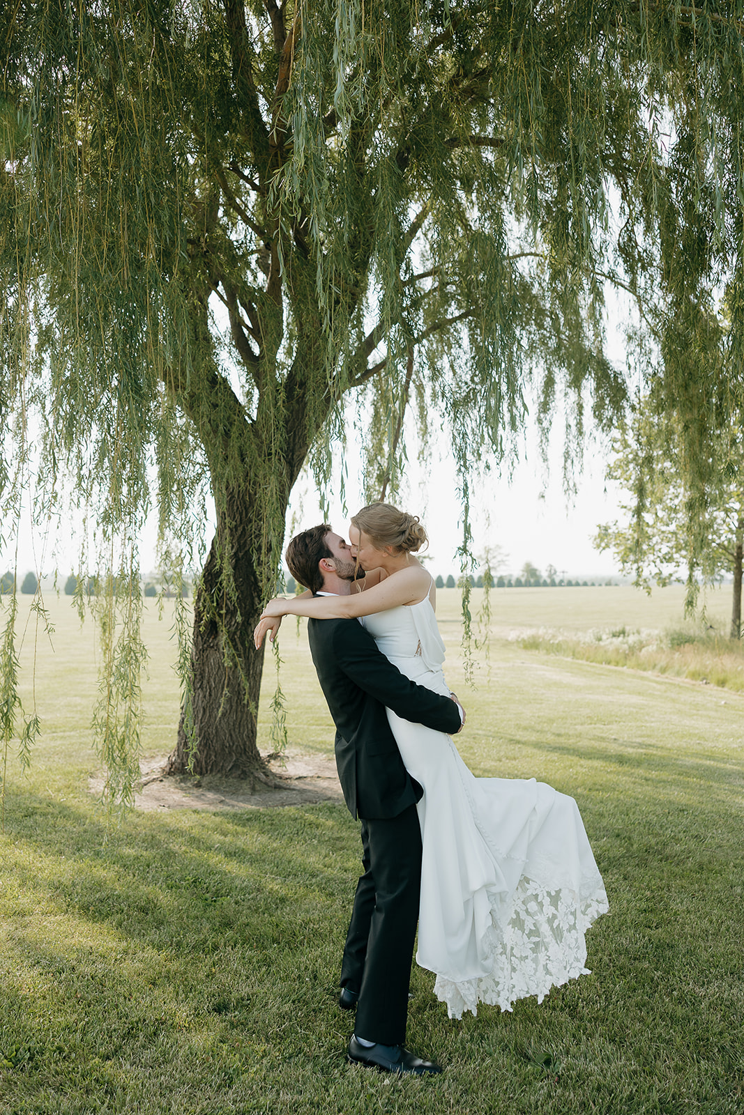 Bride and groom kissing while he lifts her off the ground beneath a willow tree at this modern wedding venue (Highpoint Event Center).