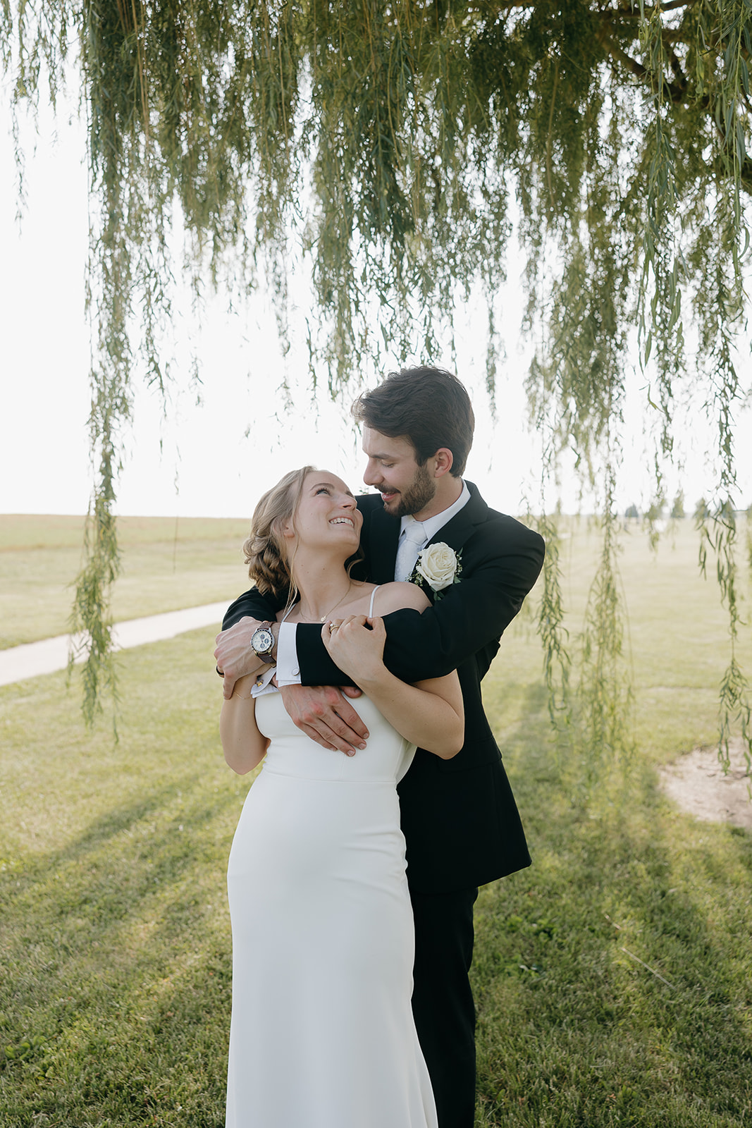 Bride smiling up at her groom while embracing under a willow tree at golden hour.