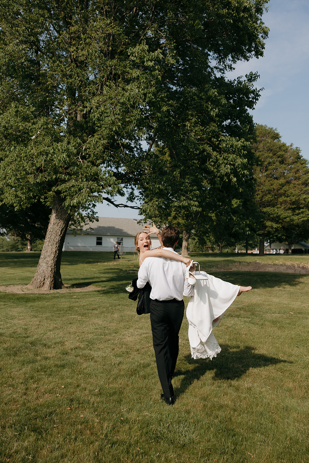 Groom carrying his laughing bride across the lawn, her bouquet and heels in hand at this modern wedding venue (Highpoint Event Center).
