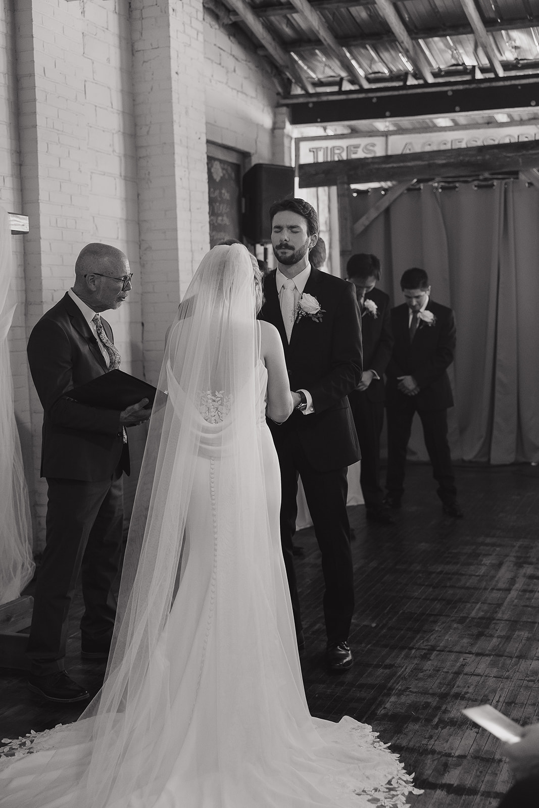 Black and white photo of the groom closing his eyes during the ceremony as the officiant prays over the wedding.