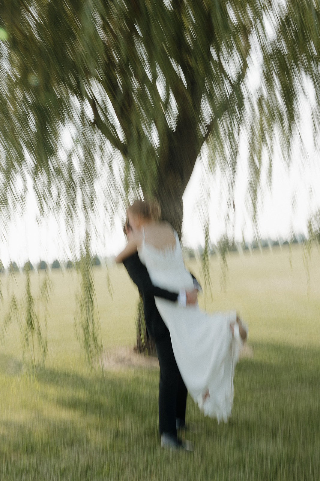 Motion blur photo of the groom lifting his bride under a weeping willow tree.