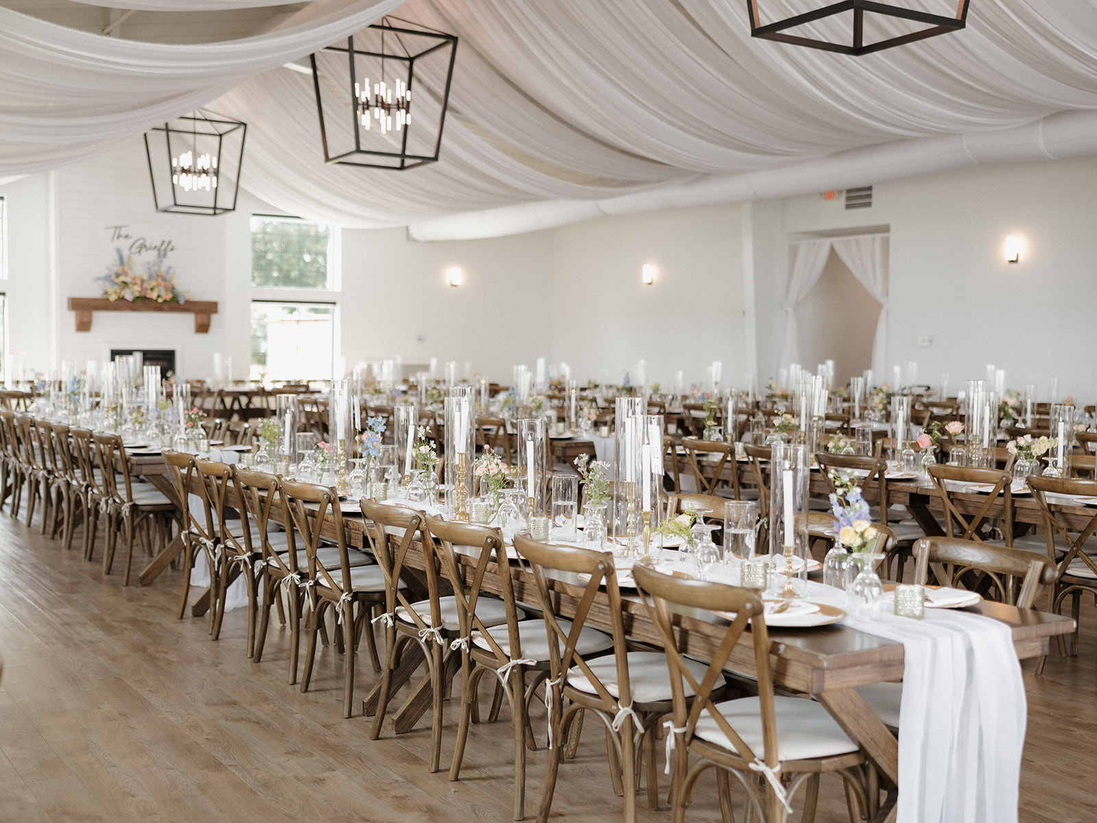 Long reception table set with gold chargers, white taper candles, glass hurricanes, and bud vases of pastel flowers, a tablescape made for a garden party wedding.
